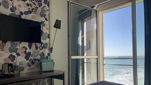 Habitación de hotel con una cama, un escritorio con lámpara, televisión y vista al mar a través de un ventanal grande.