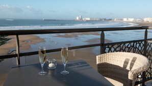Un balcón con vistas despejadas a la playa y al océano, una mesa con dos copas de champán y una vela, y una silla de mimbre.