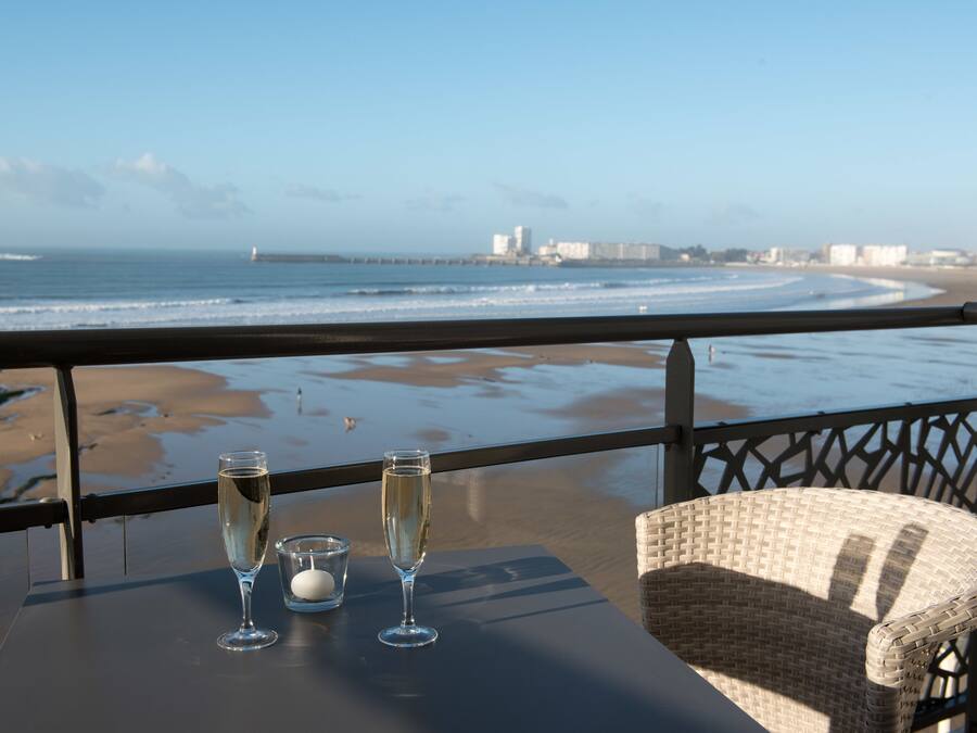 Un balcon offrant une vue dégagée sur la plage et l’océan, une table avec deux coupes à champagne et une bougie, et un fauteuil en osier.