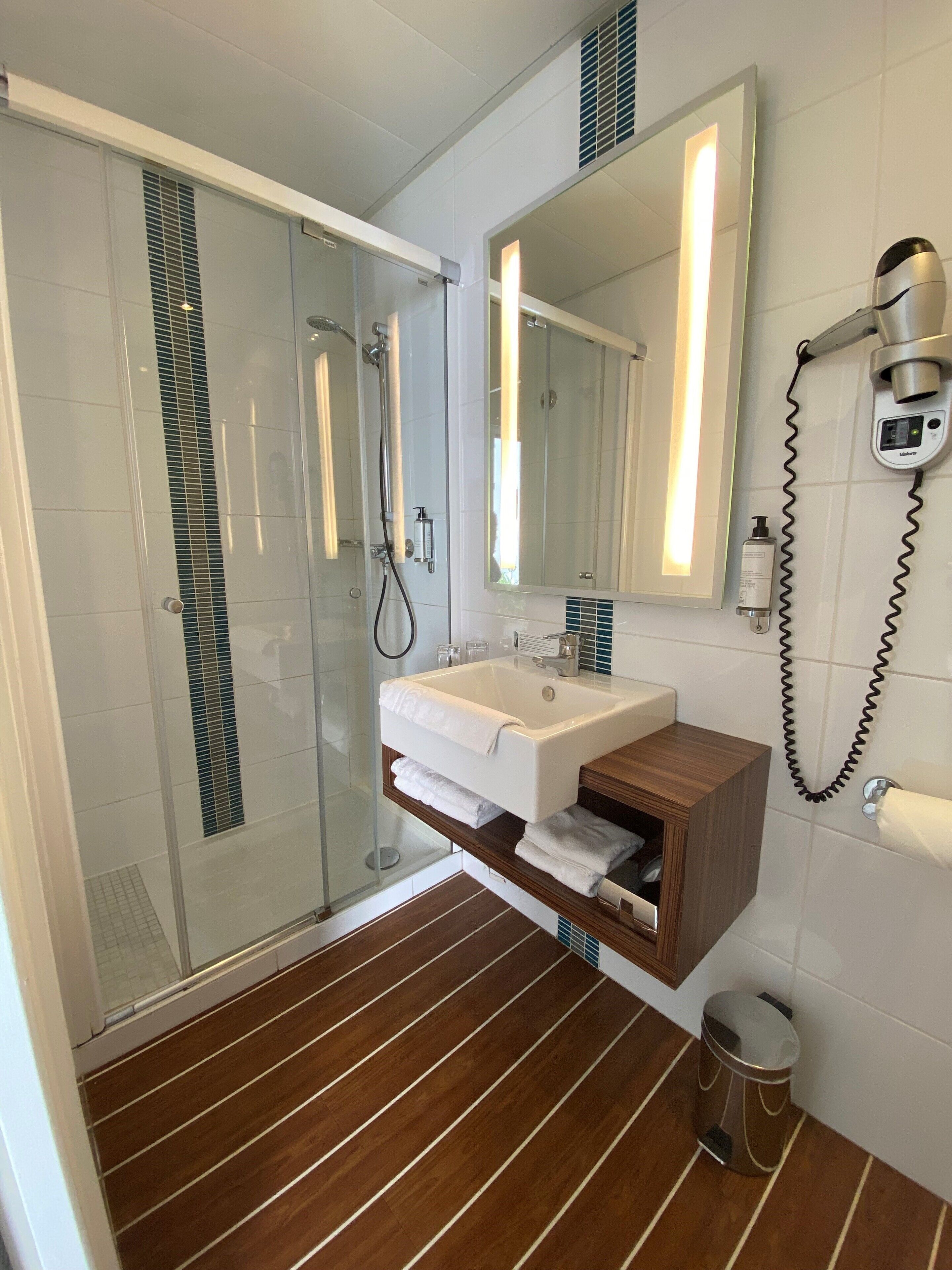 Un baño moderno con cabina de ducha de vidrio, un lavabo blanco sobre un mueble de madera, un espejo grande y un secador de pelo en la pared.