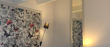 A hotel room with a bed, a desk with a lamp, a mirror, and a floral wallpaper.