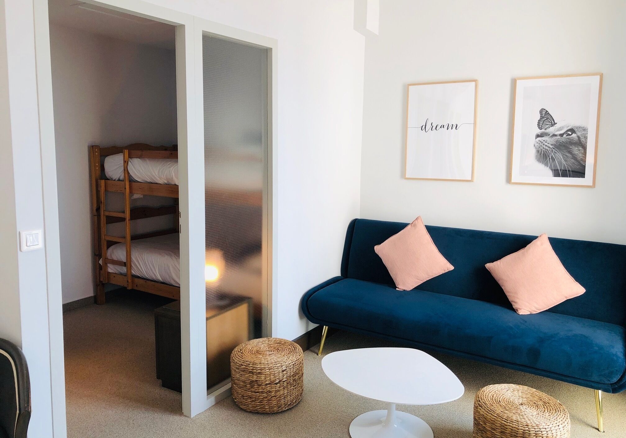 Una habitación con una cama litera, un sofá azul con dos cojines, una mesa de centro blanca y dos taburetes de mimbre. Hay dos cuadros enmarcados en la pared.