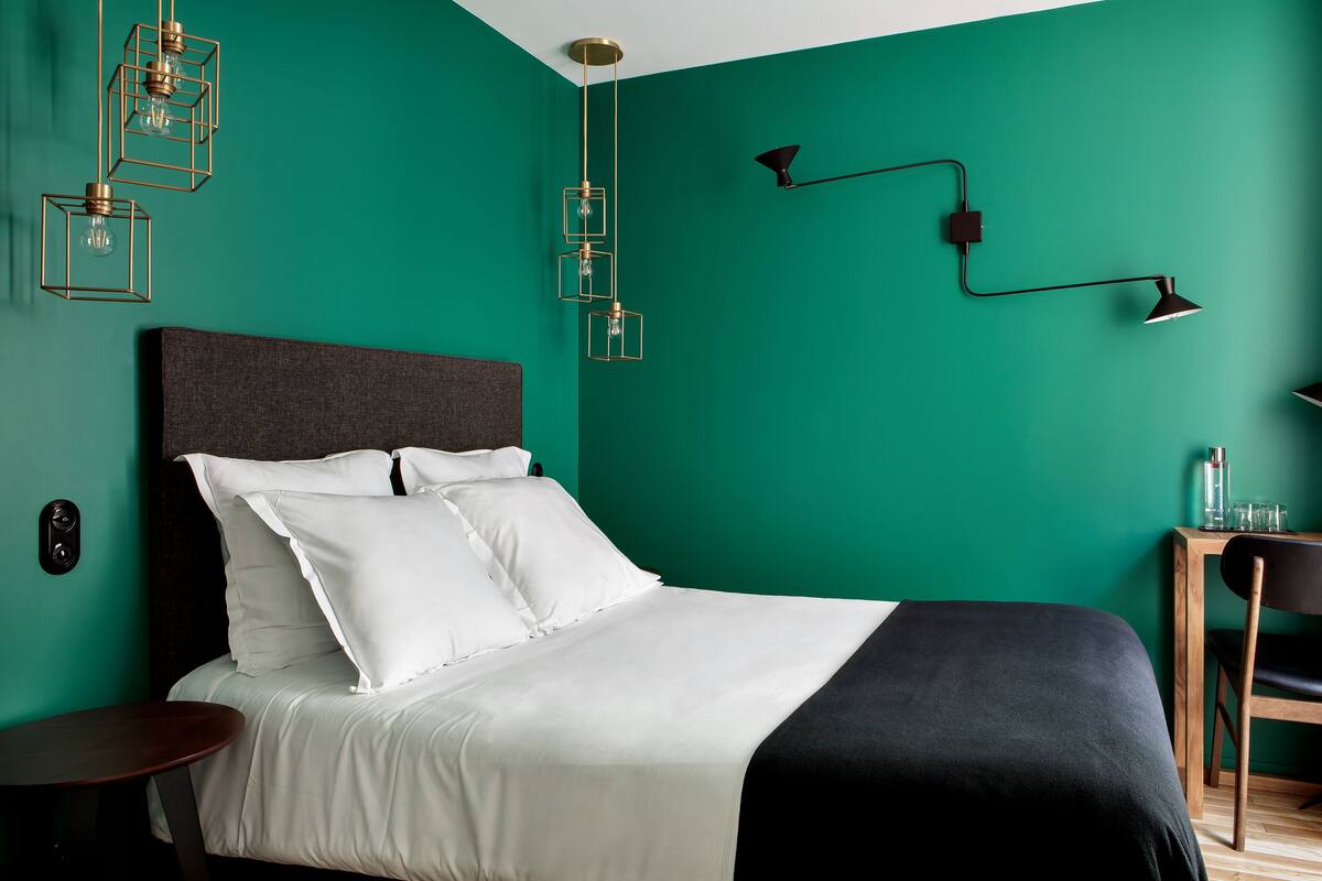 A neatly made bed with white and black bedding, a dark headboard, and a teal wall. There are three hanging light fixtures and a wall-mounted lamp.