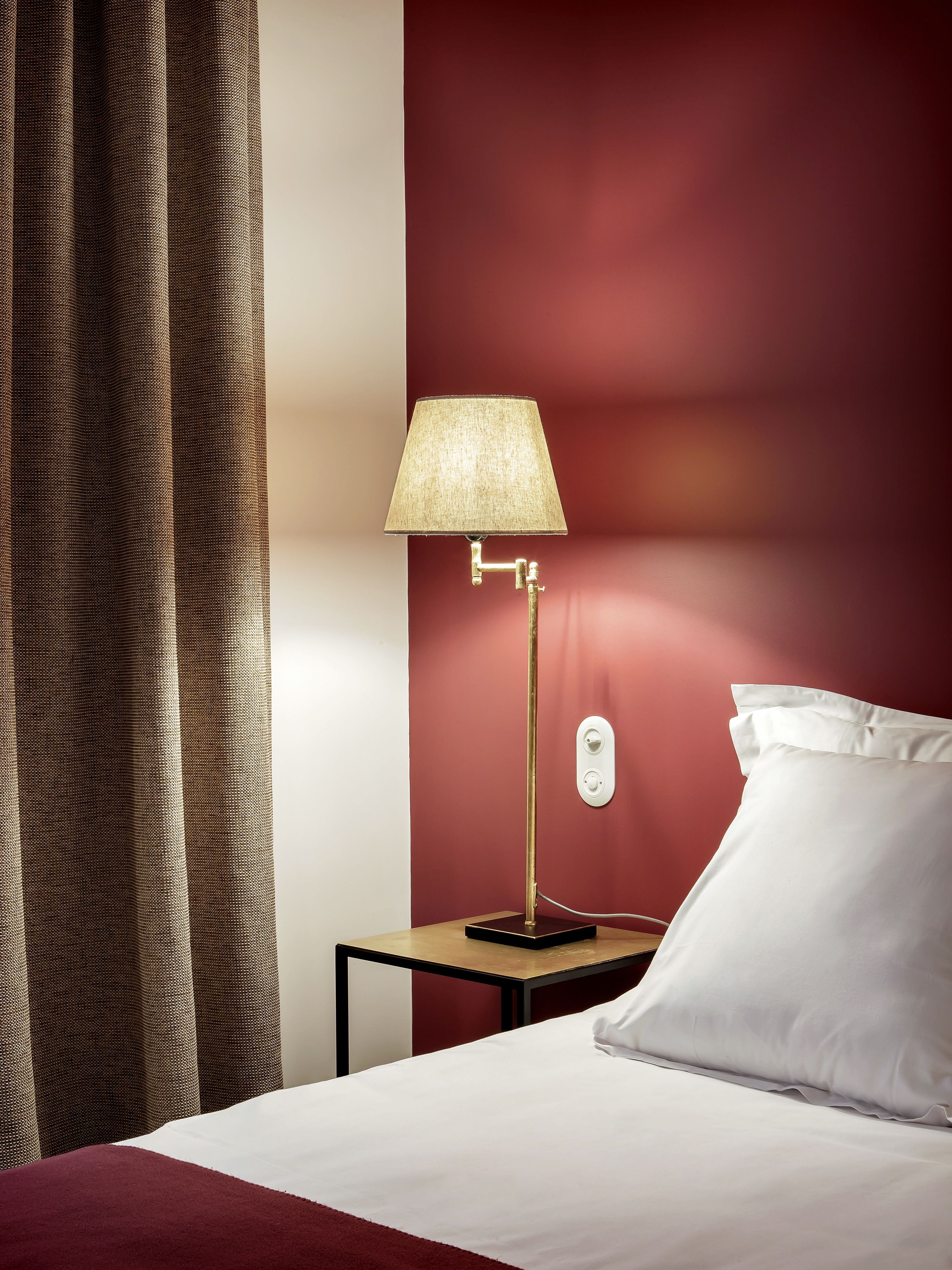 A neatly made bed with white linens, a gold lamp on a small table beside it, and a red wall in the background.