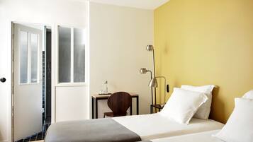 Une chambre d’hôtel avec deux lits, un bureau, une chaise, une lampe et une vue sur un espace carrelé, visible à travers une porte ouverte.