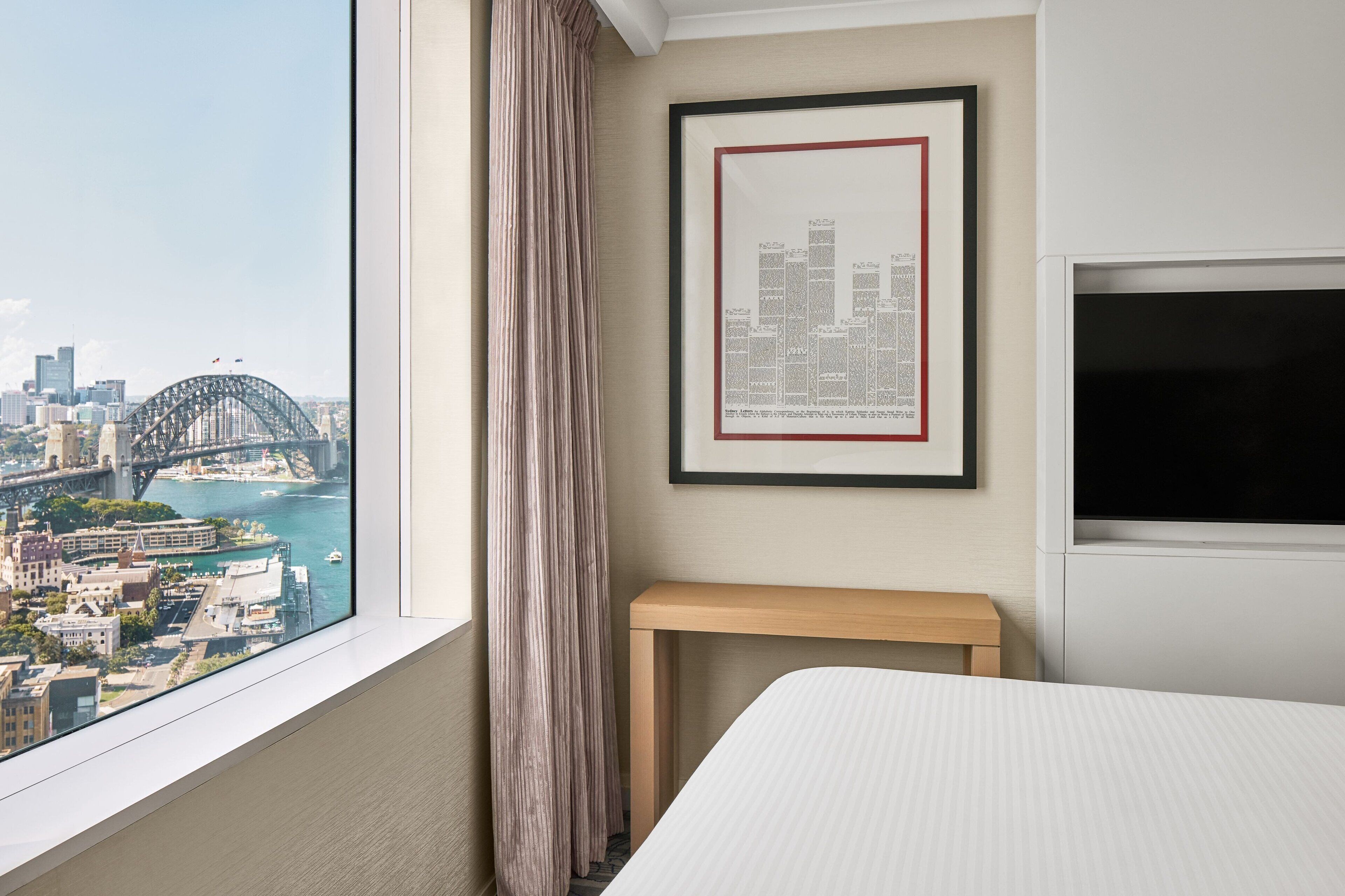 A hotel room with a large window offering a cityscape view, a framed city skyline artwork, a wooden bedside table, and a bed.
