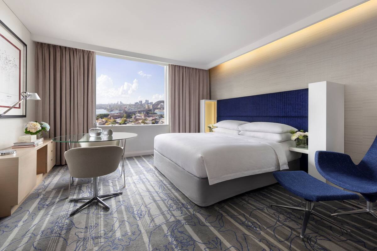 A modern hotel room with a large bed, a desk, a chair, and a view of a cityscape.