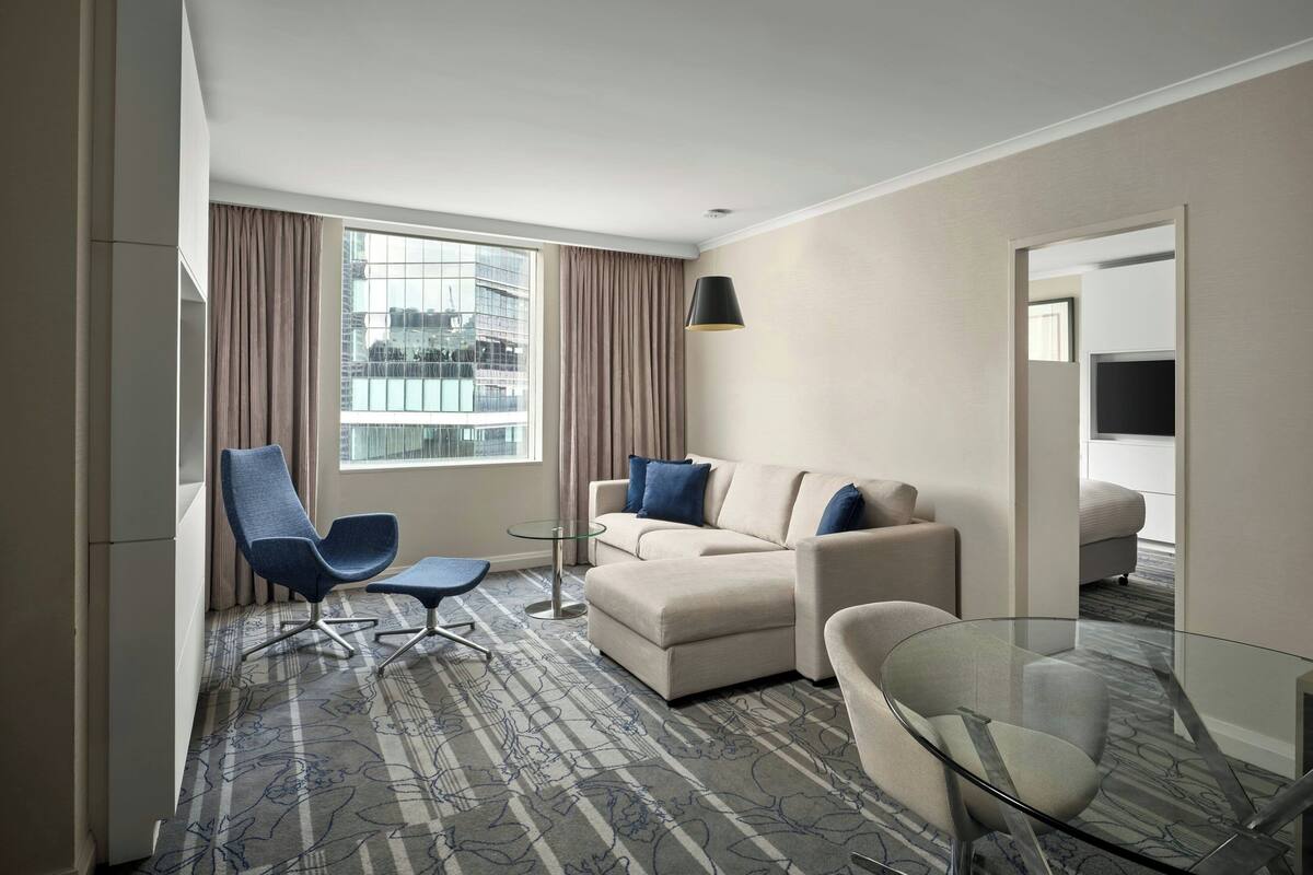 A modern hotel room with a large window, a sofa, a glass table, and a chair.