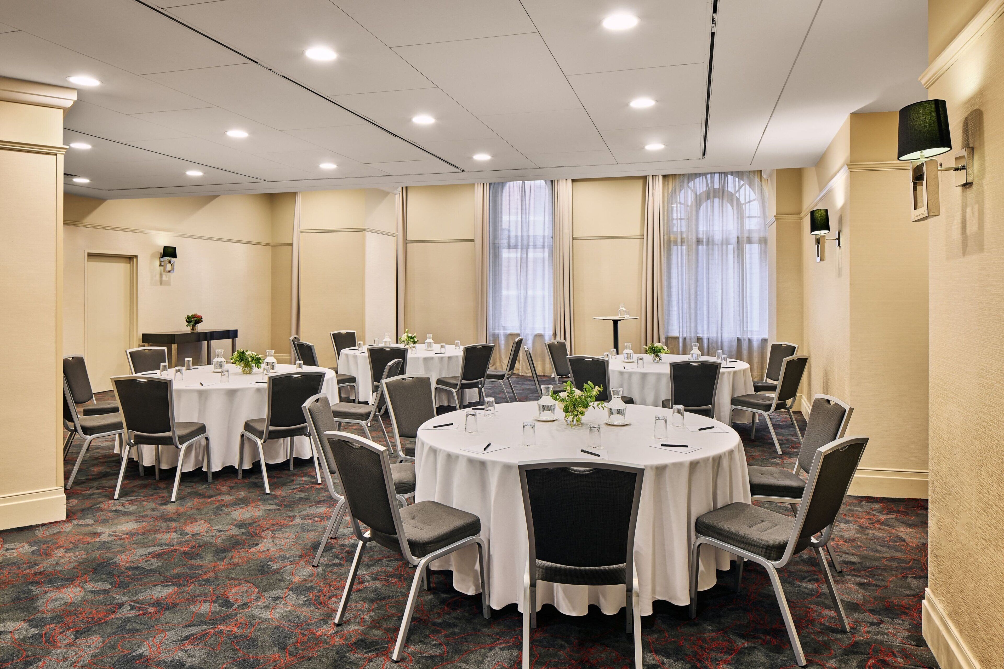 Un salón de conferencias con varias mesas redondas, cada una rodeada de sillas, y equipado con botellas de agua y pequeñas plantas.