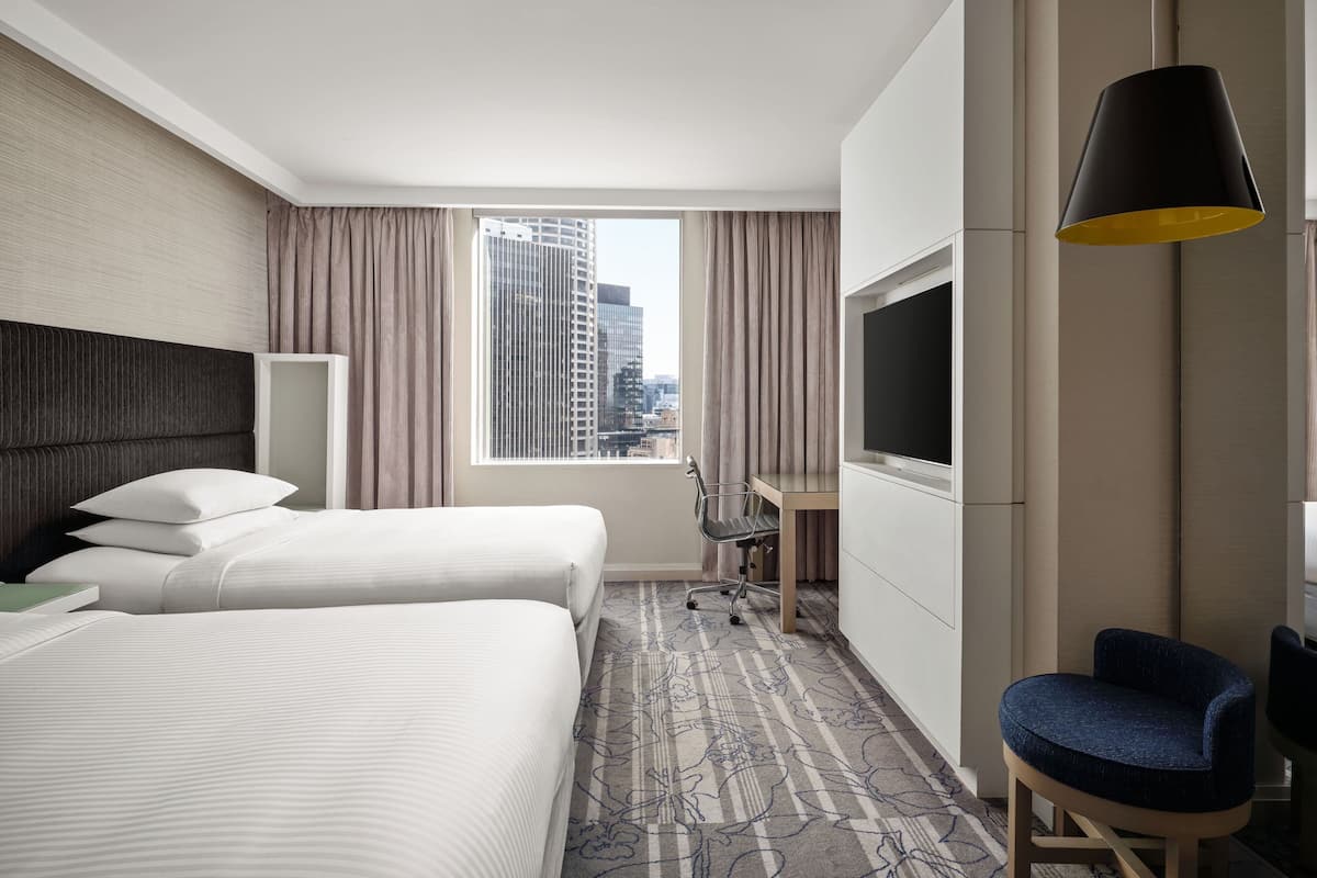 A hotel room with two beds, a desk, a chair, a TV, and a view of the cityscape.