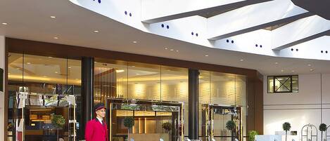 A modern hotel entrance with a glass facade, a reception area, and a staff member in uniform.