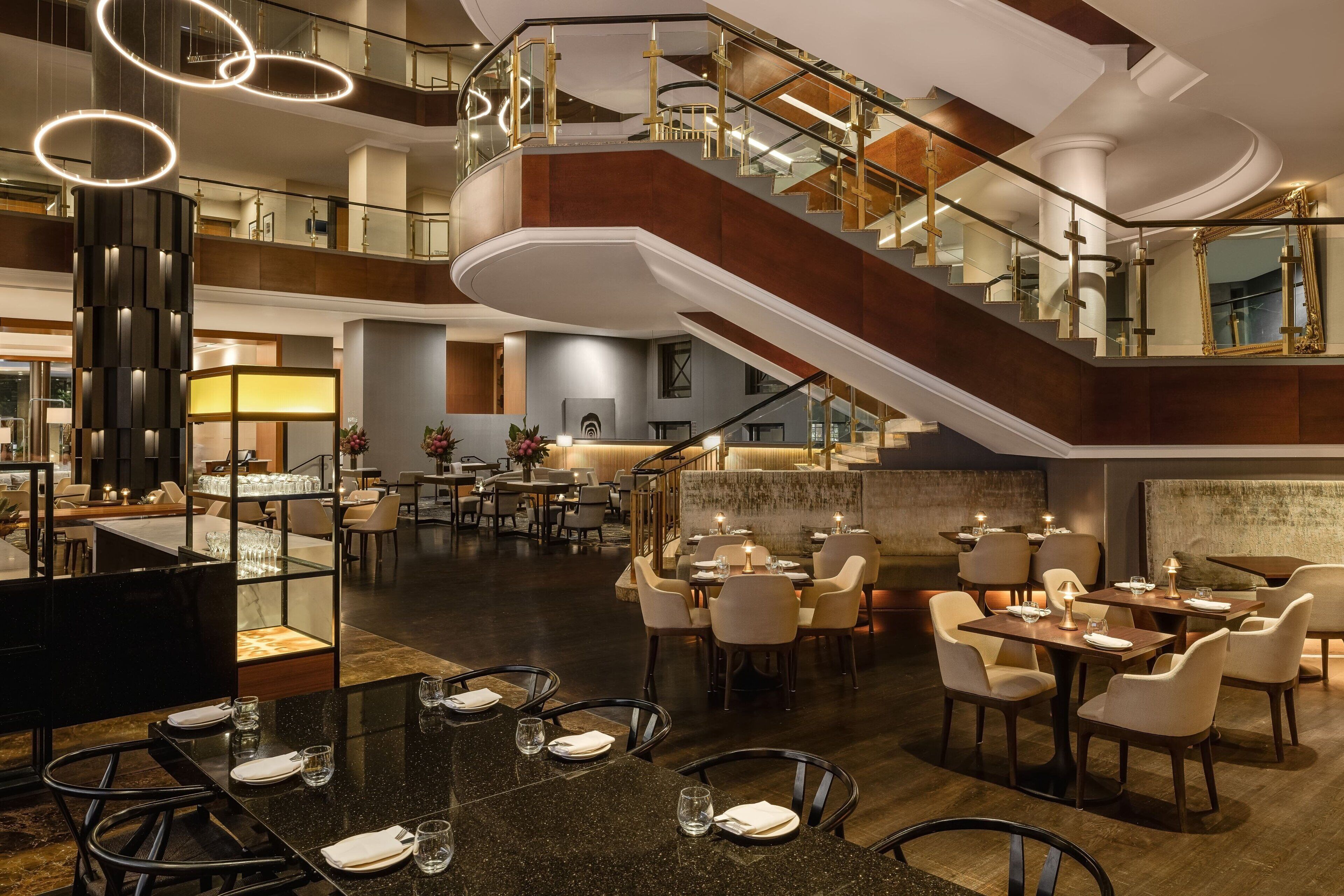 A spacious hotel lobby with a grand staircase, multiple dining areas, and a modern chandelier.