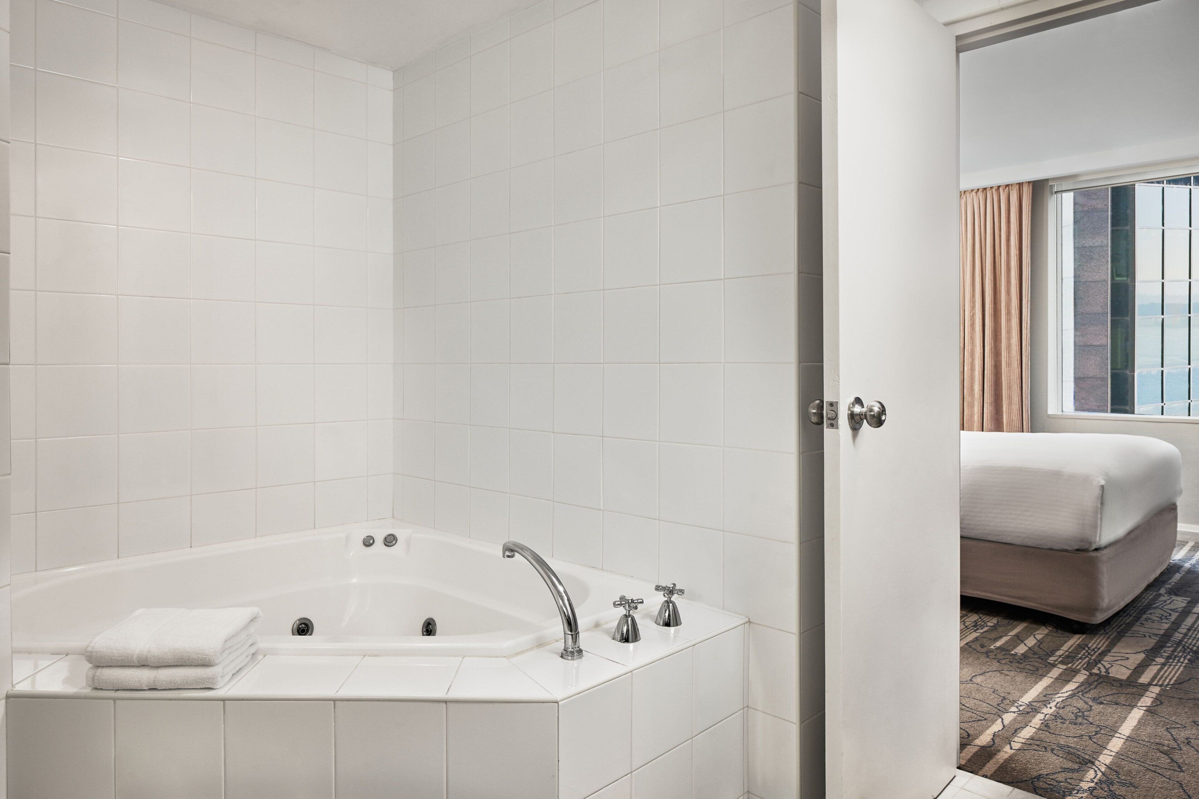 A modern bathroom with a large bathtub, a toilet, and a sink. There is a bed with a window and curtains in the adjacent room.