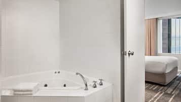 A modern bathroom with a large bathtub, a toilet, and a sink. There is a bed with a window and curtains in the adjacent room.