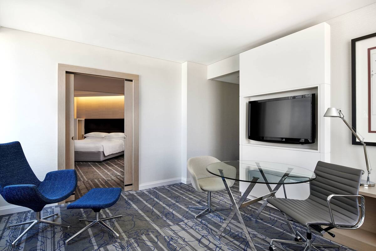 A hotel room with a bed, two blue chairs, a glass table, a TV, and a lamp.