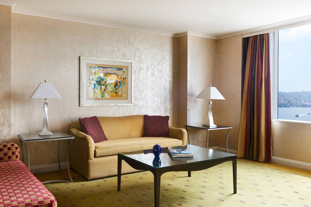 A hotel room with a sofa, a coffee table, two lamps, a painting, and a window with curtains.