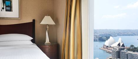A hotel room with a bed, a nightstand, a lamp, and a view of the Sydney Opera House and harbor.