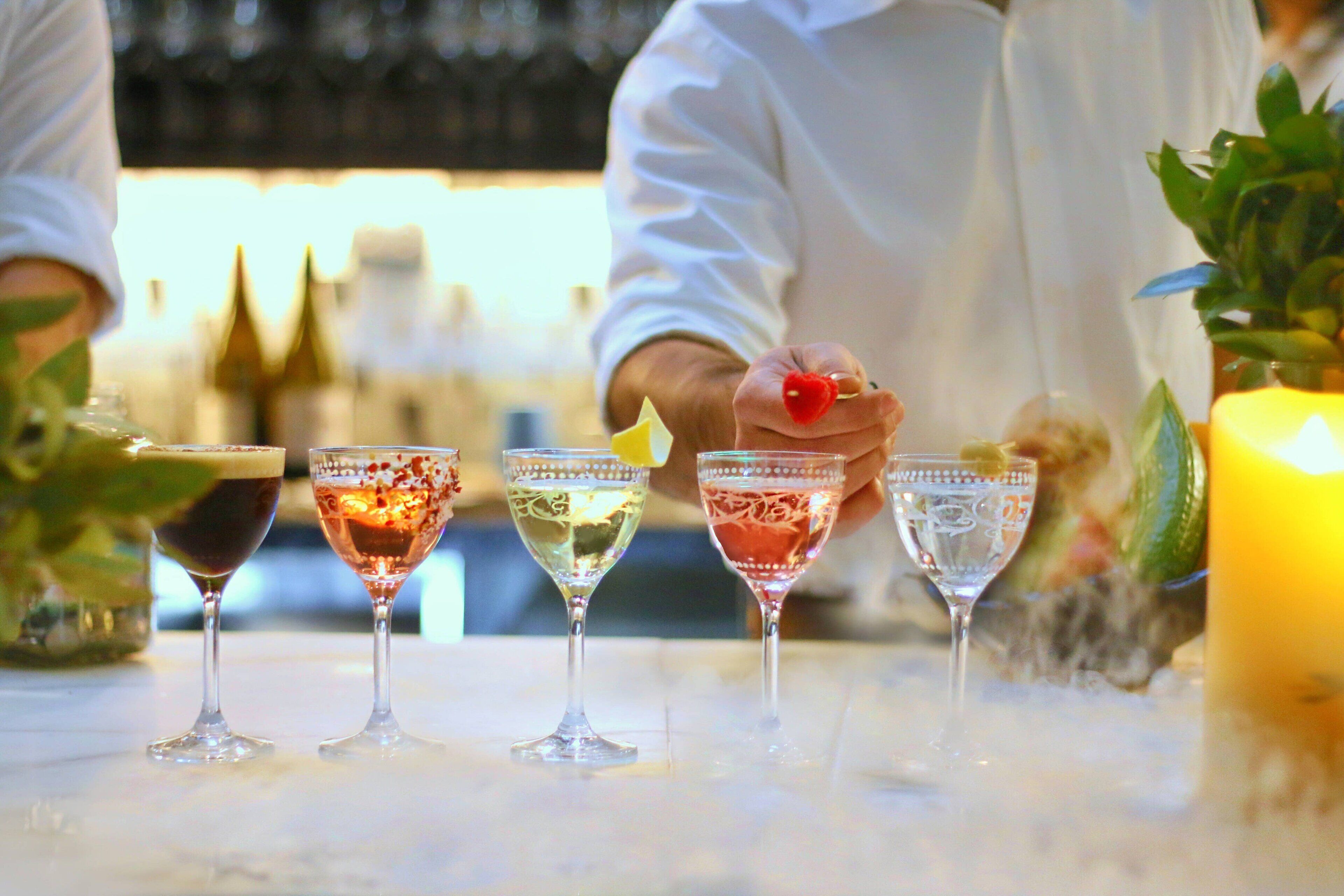 A bar with four glasses of various cocktails, a candle, and a potted plant.