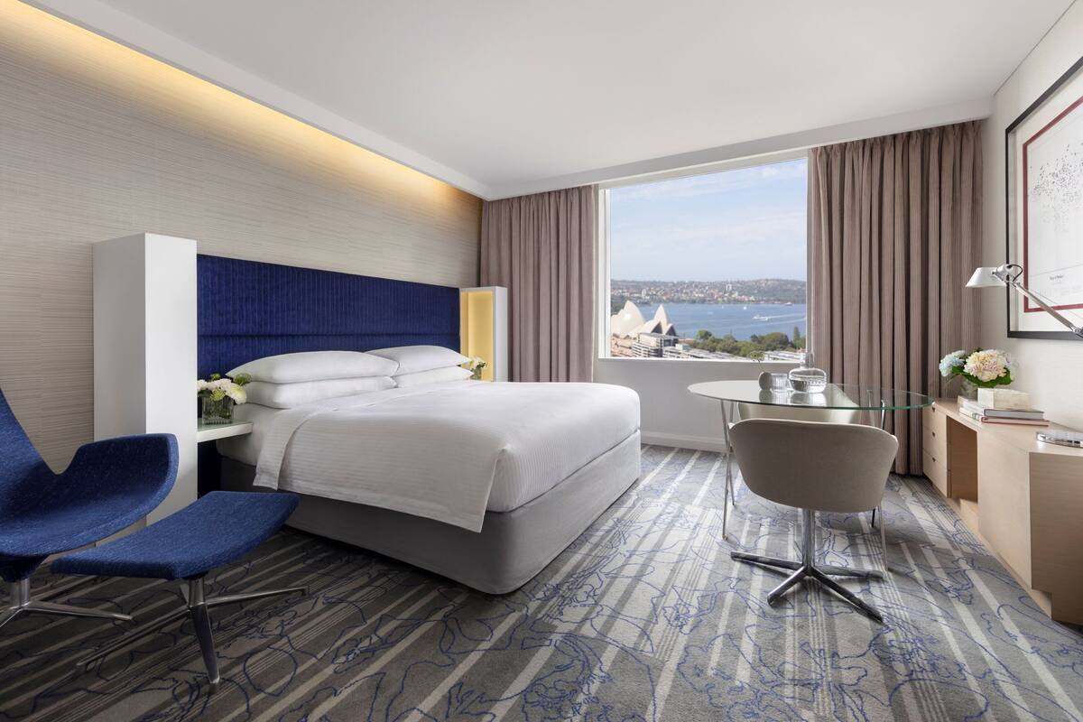A hotel room with a bed, a desk, a chair, a small table, and a view of a cityscape.