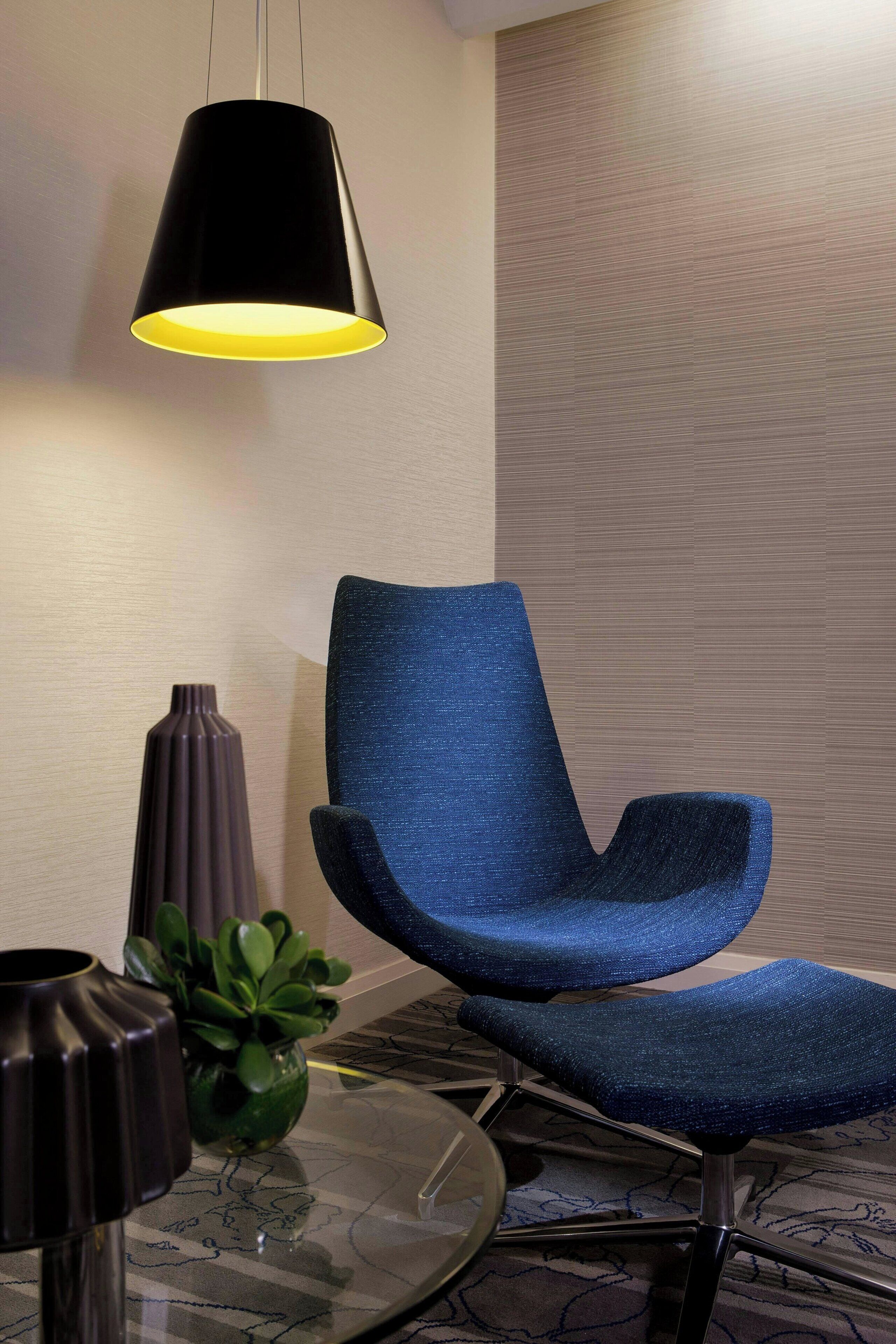 A modern room with a blue upholstered chair, a black pendant light, a glass table, and a green plant.