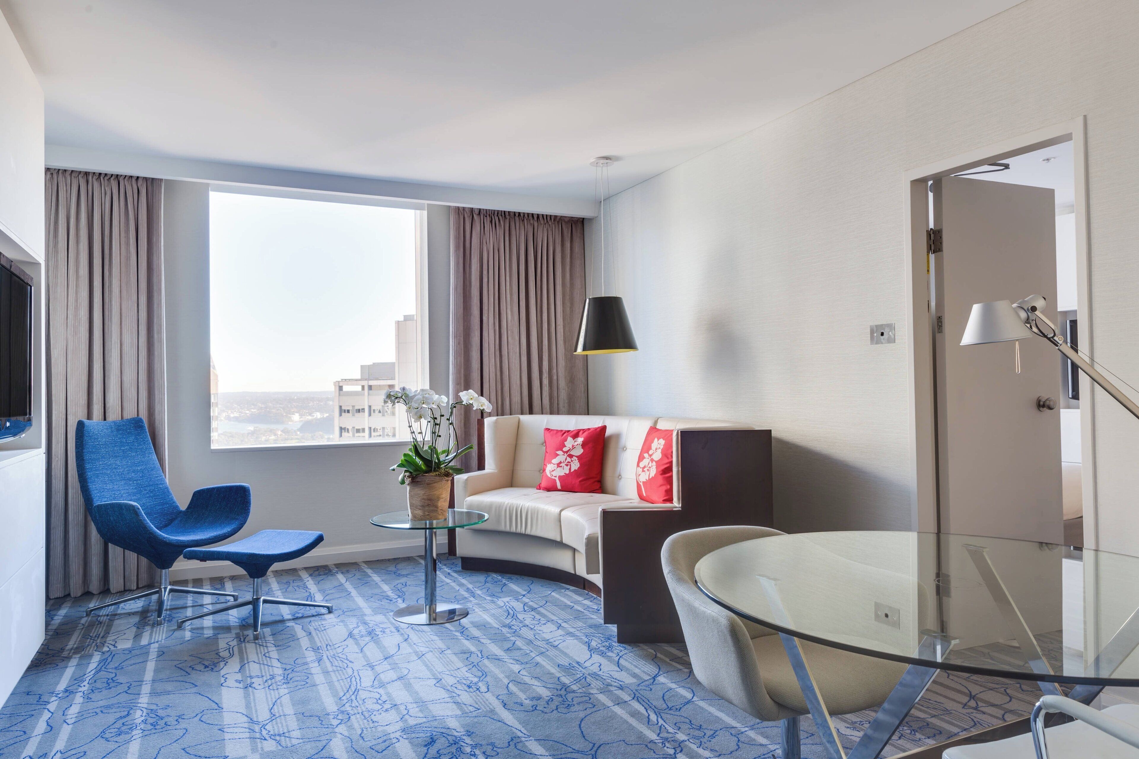 Habitación de hotel moderna con un sofá curvo, un sillón azul, una mesa de vidrio y un ventanal con vistas a la ciudad.