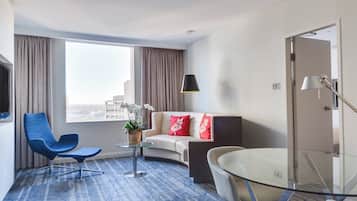 Habitación de hotel moderna con un sofá curvo, un sillón azul, una mesa de vidrio y un ventanal con vistas a la ciudad.