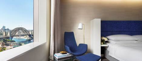 A hotel room with a view of a bridge, a bed, a blue chair, a small table with books, and a lamp.