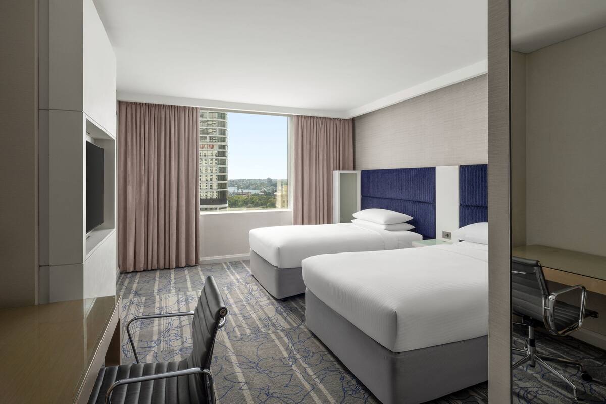 A hotel room with two beds, a large window with curtains, a flat-screen TV, and a seating area with a chair and a table.