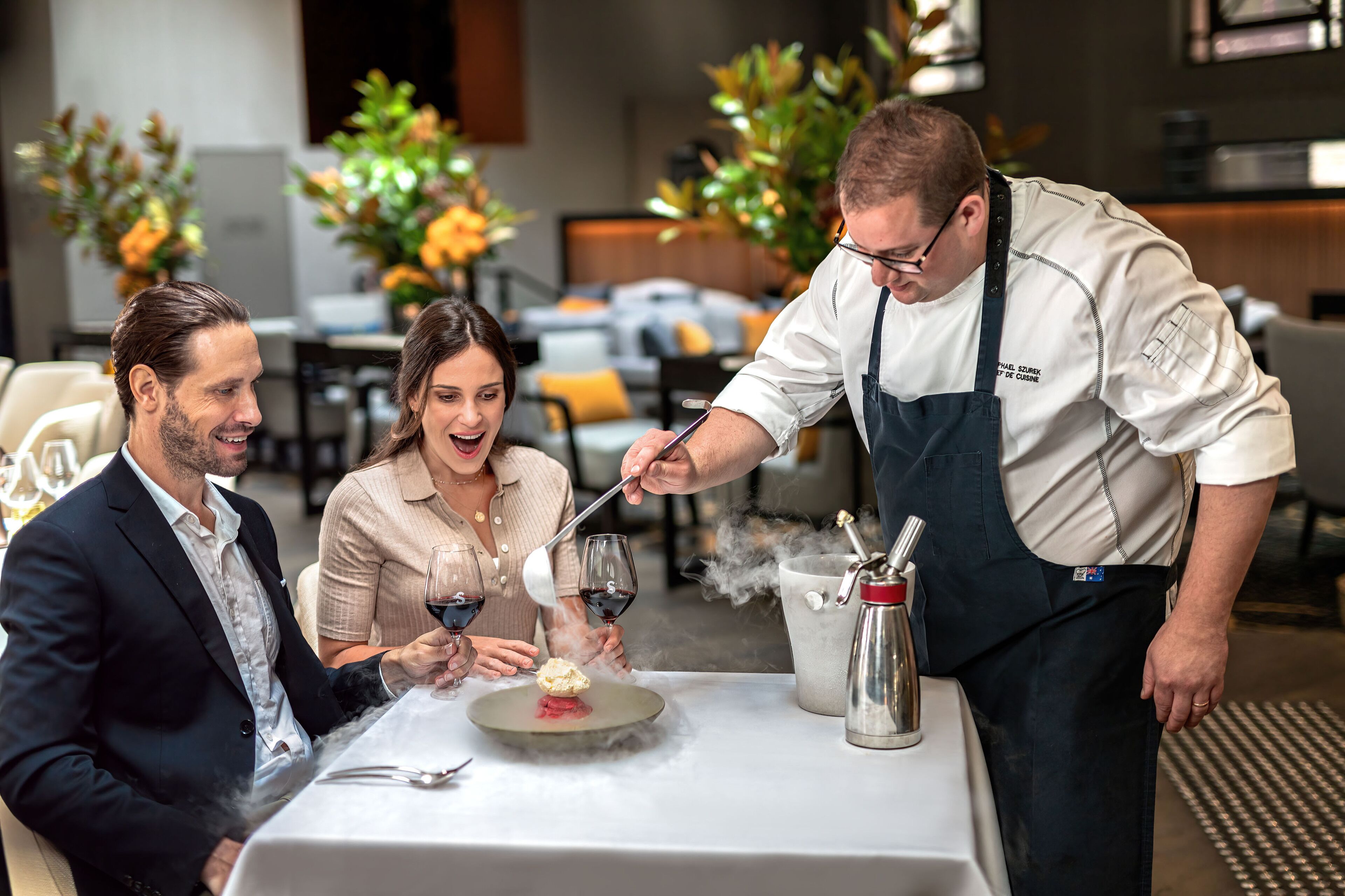 A dining table with a dessert being served, a waiter in uniform, and a couple seated.
