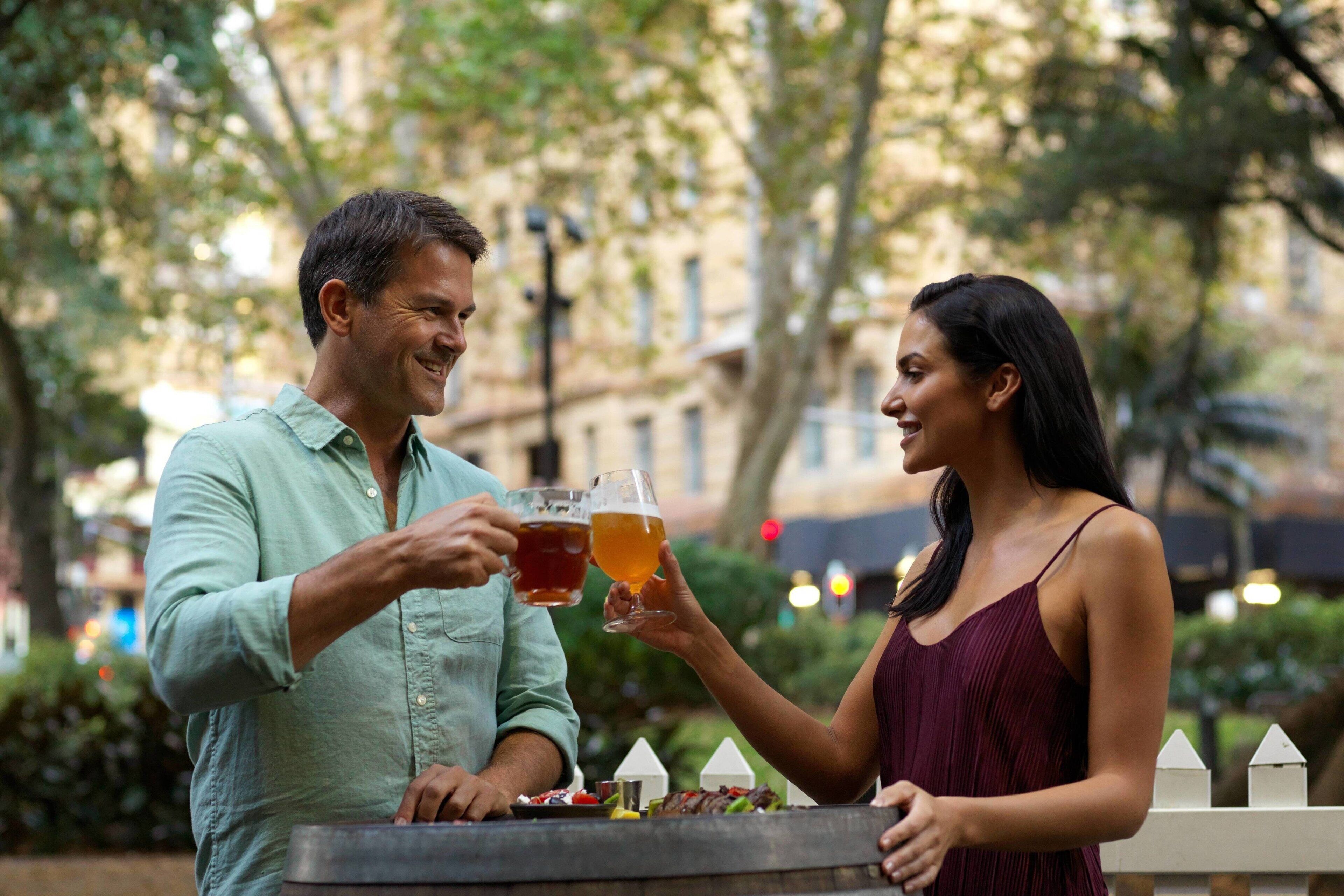 A couple toasting with drinks at an outdoor table with a view of buildings and trees.