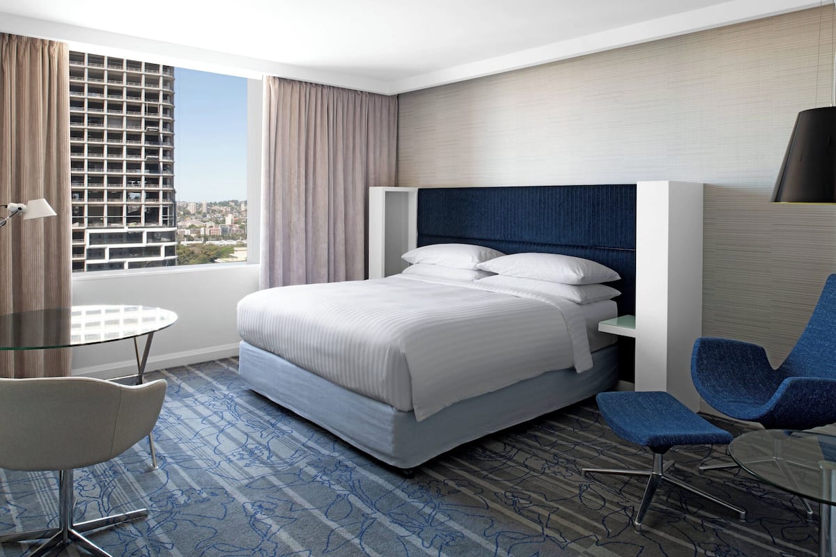 A hotel room with a large bed, a desk, a chair, and a view of a cityscape through the window.