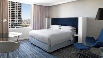 Habitación de hotel con una cama grande, un escritorio, una silla y vista a un paisaje urbano a través de la ventana.
