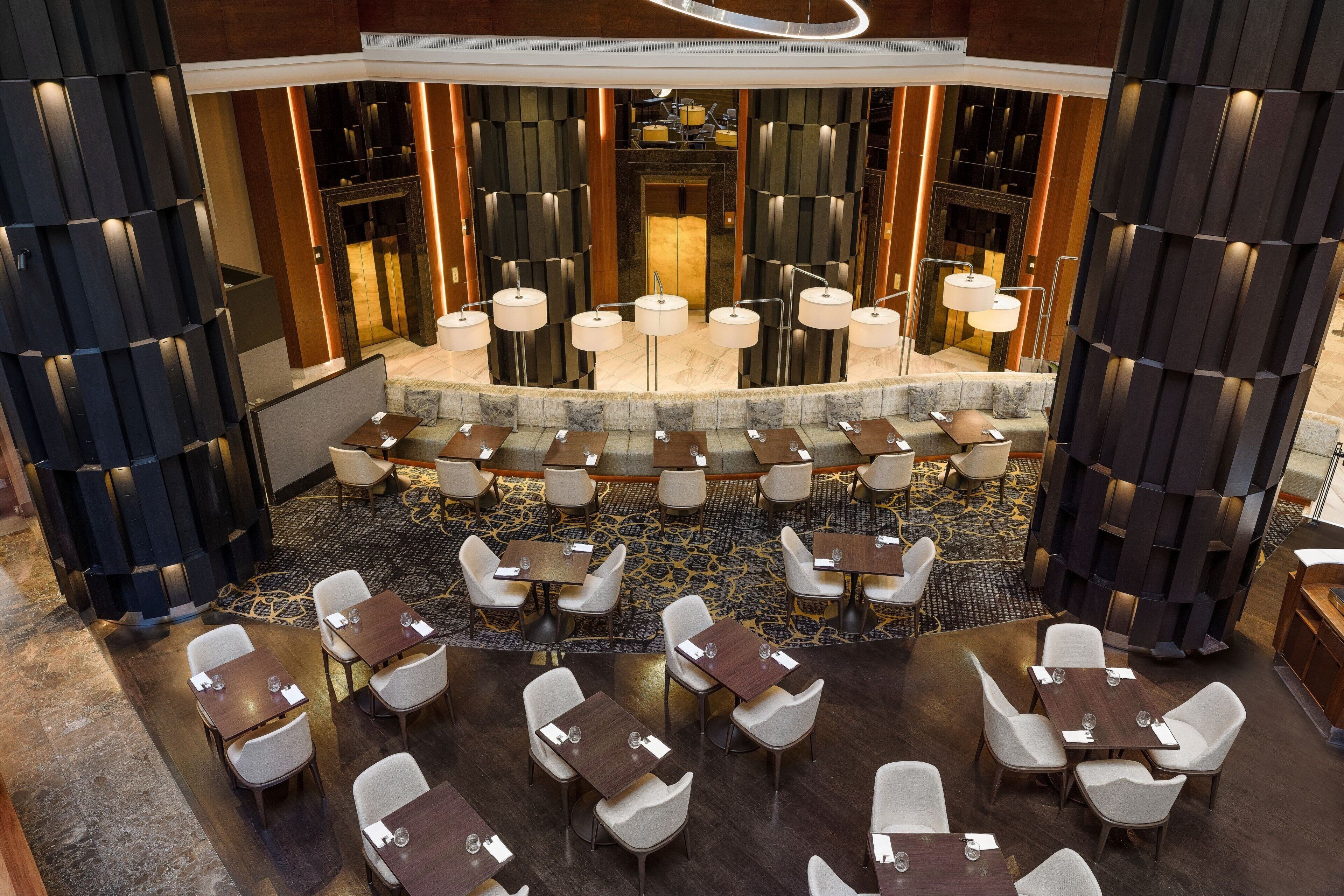 A modern hotel lobby with a central seating area, multiple tables, and a patterned carpet.