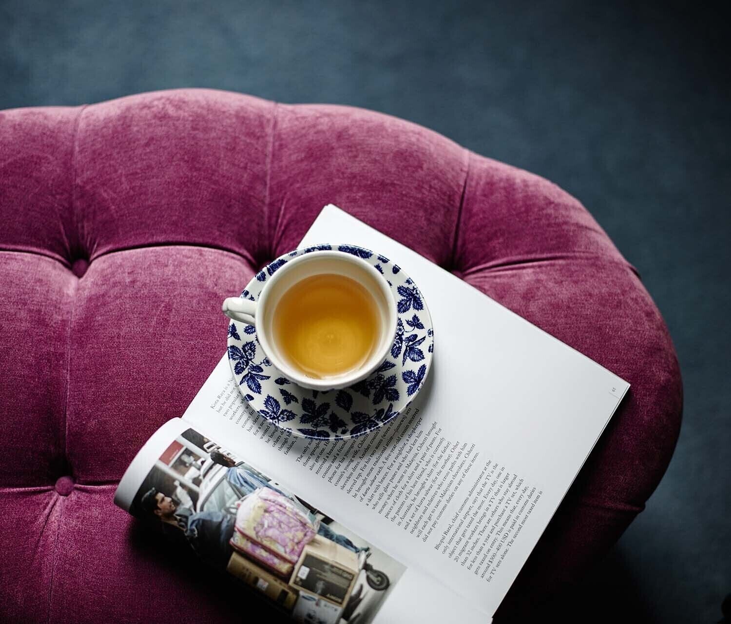 Una taza de té sobre un plato con diseño, una revista con una foto de una persona y un cojín.