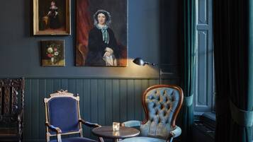 A room with a dark blue wall, a large portrait, a wooden chair, a smaller chair, a round table with a candle, and a window with curtains.