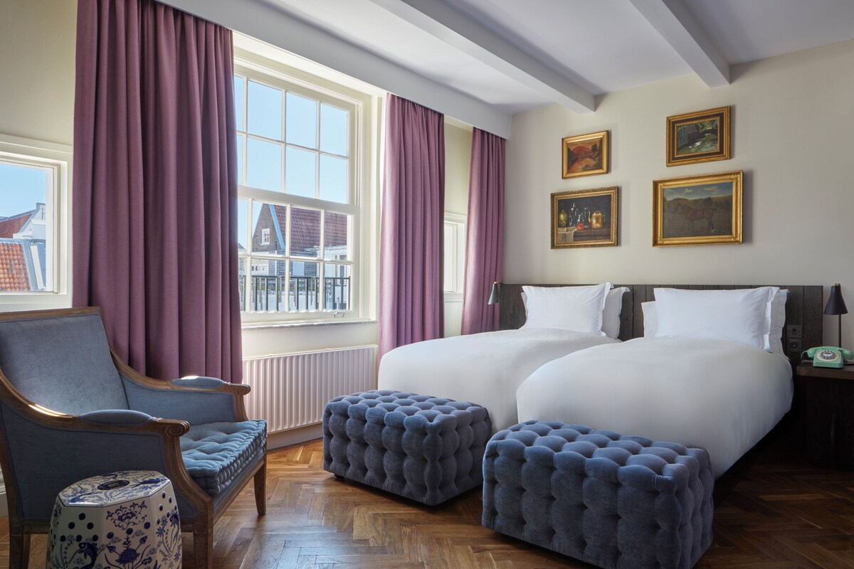 Habitación de hotel con una cama grande, dos mesitas de noche, una silla y una ventana con cortinas.