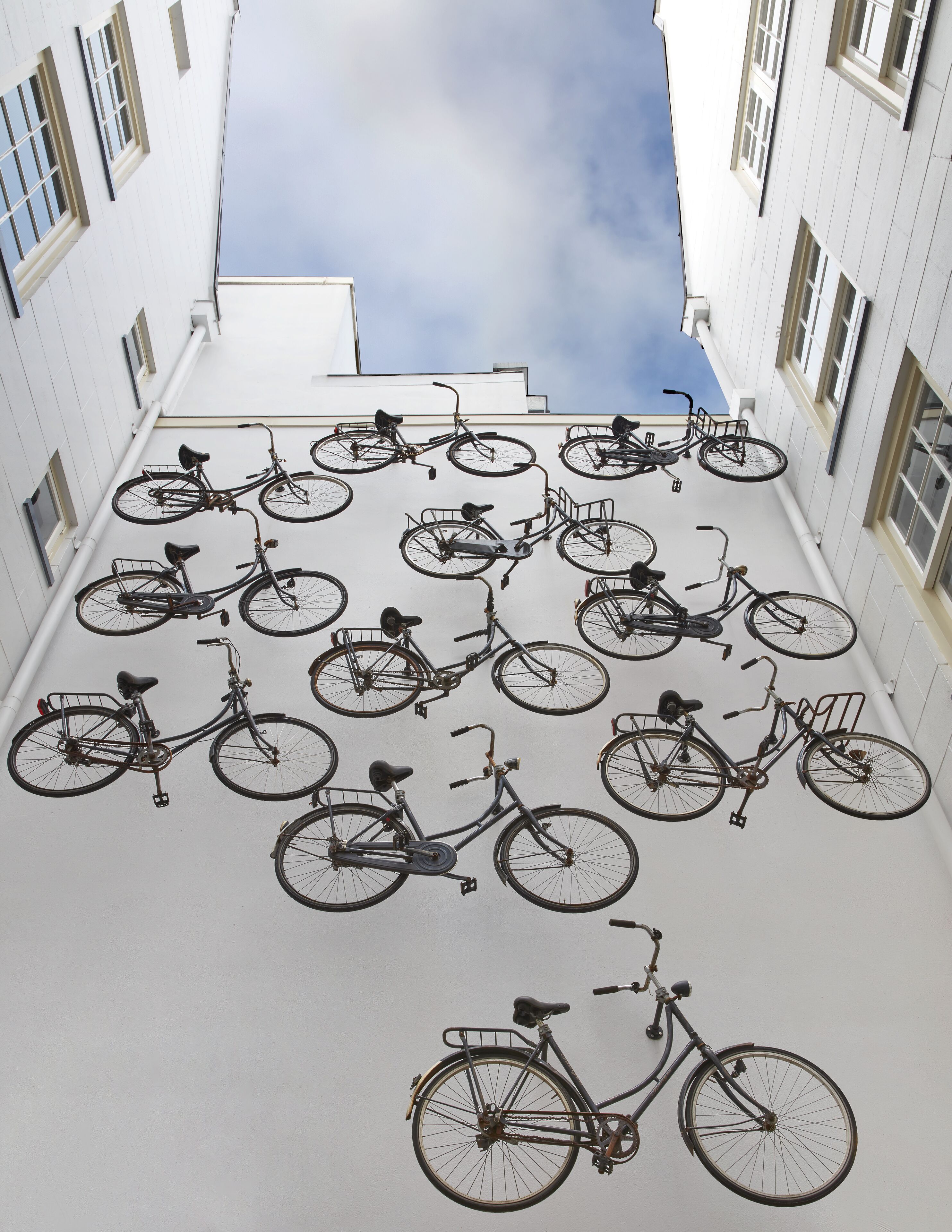 A courtyard with multiple bicycles parked against a white wall, surrounded by buildings with windows and a clear sky above.