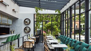 Un espacio de comedor al aire libre con mesas y sillas, una zona de bar con taburetes y grandes ventanales con follaje exterior.