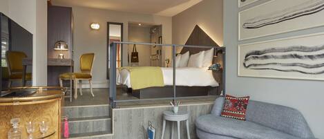 A modern hotel room with a bed, a sofa, a small table, a mirror, and a bar area with glasses.