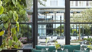 Un restaurante con asientos tapizados en verde, sillas negras y un amplio ventanal con vistas a edificios y árboles.