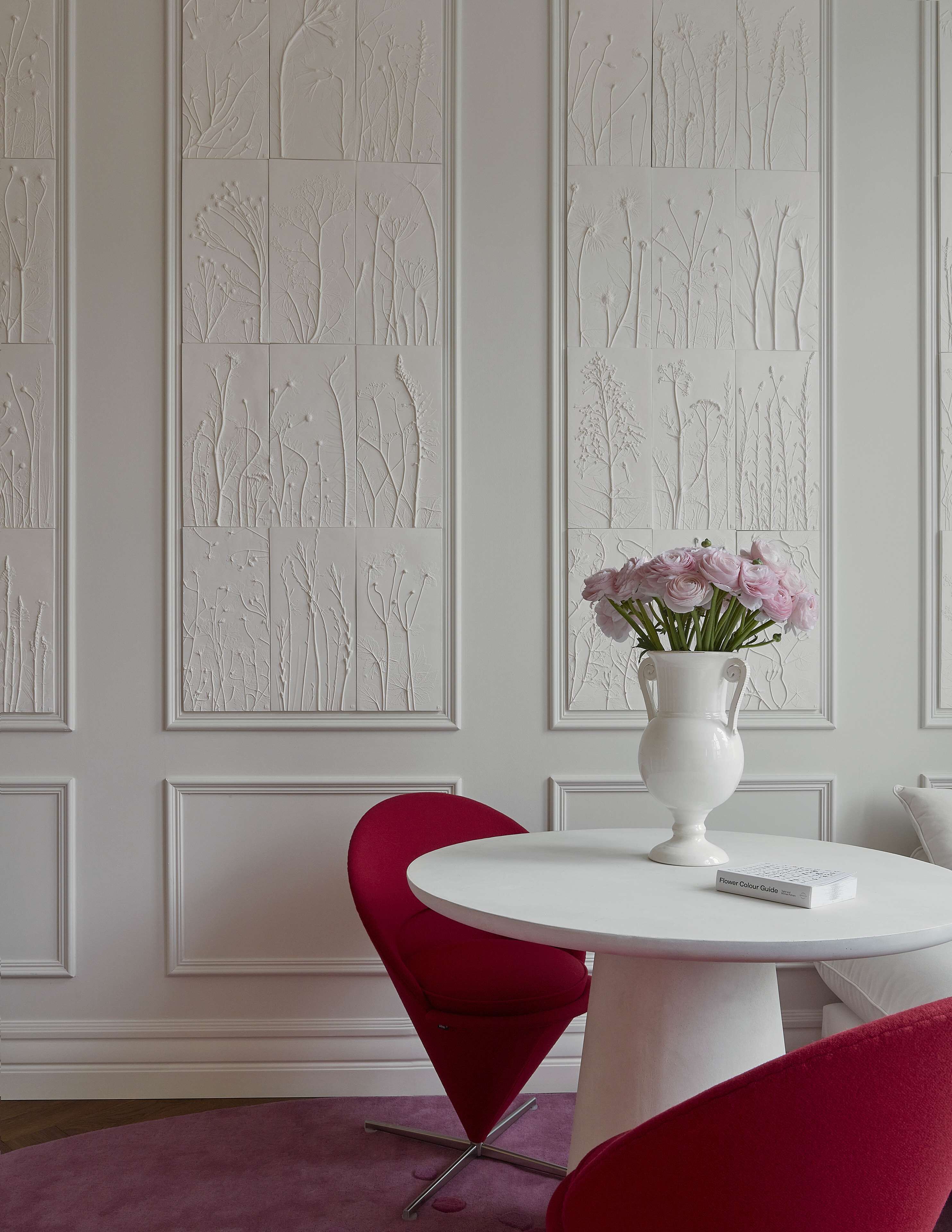 Une pièce avec un mur blanc orné de motifs floraux et d’arbres en relief, une table blanche sur laquelle est posé un vase rempli de fleurs roses, et une chaise rouge.