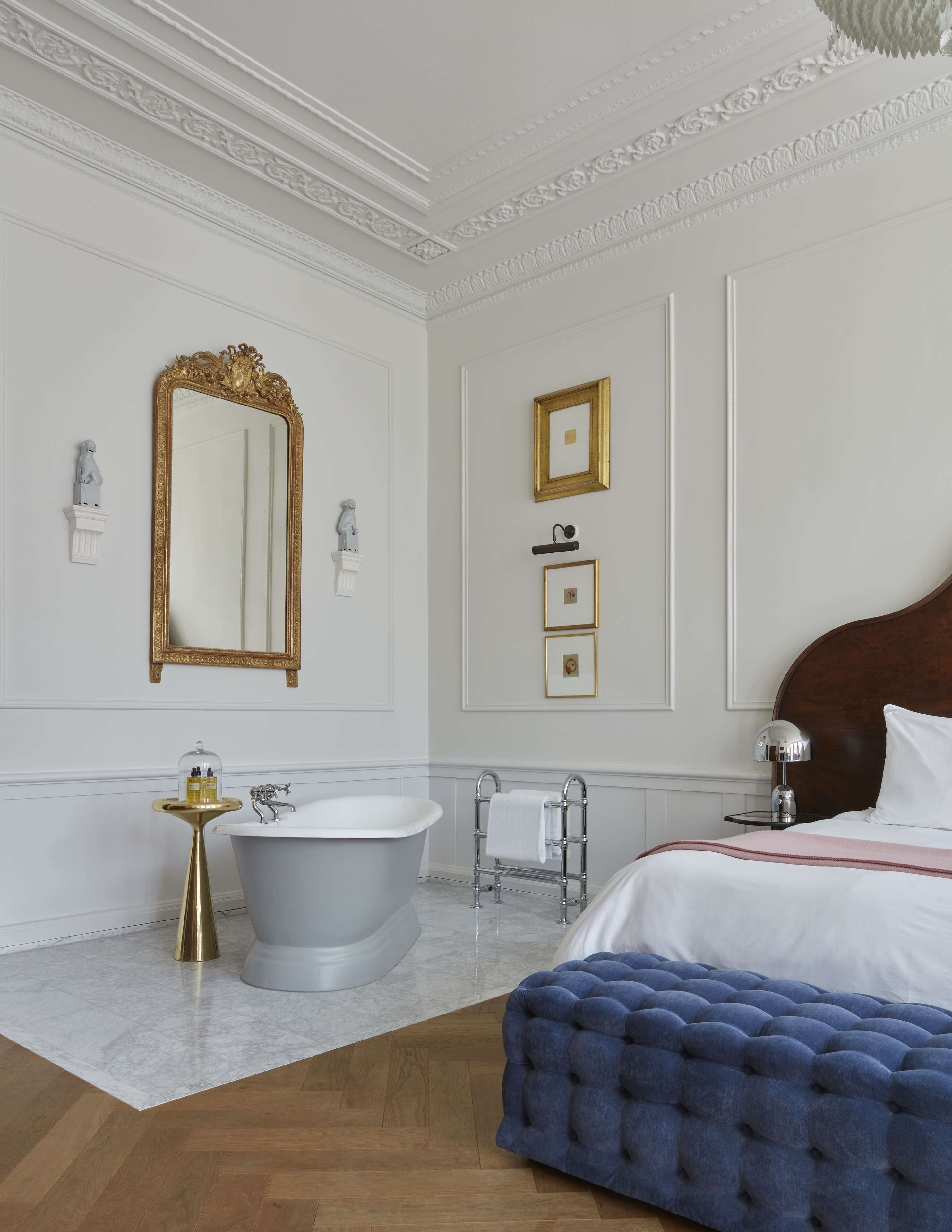 Un dormitorio lujoso con una cama grande, una bañera independiente, un espejo con marco dorado y un piso de mármol.
