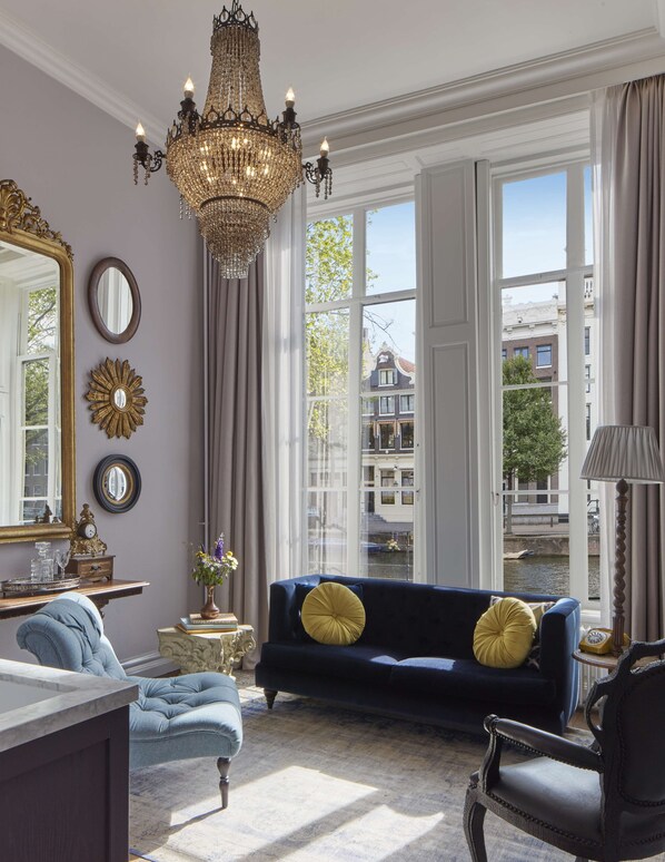 A room with a large chandelier, a blue sofa with yellow pillows, a mirror, and a view of buildings through large windows.