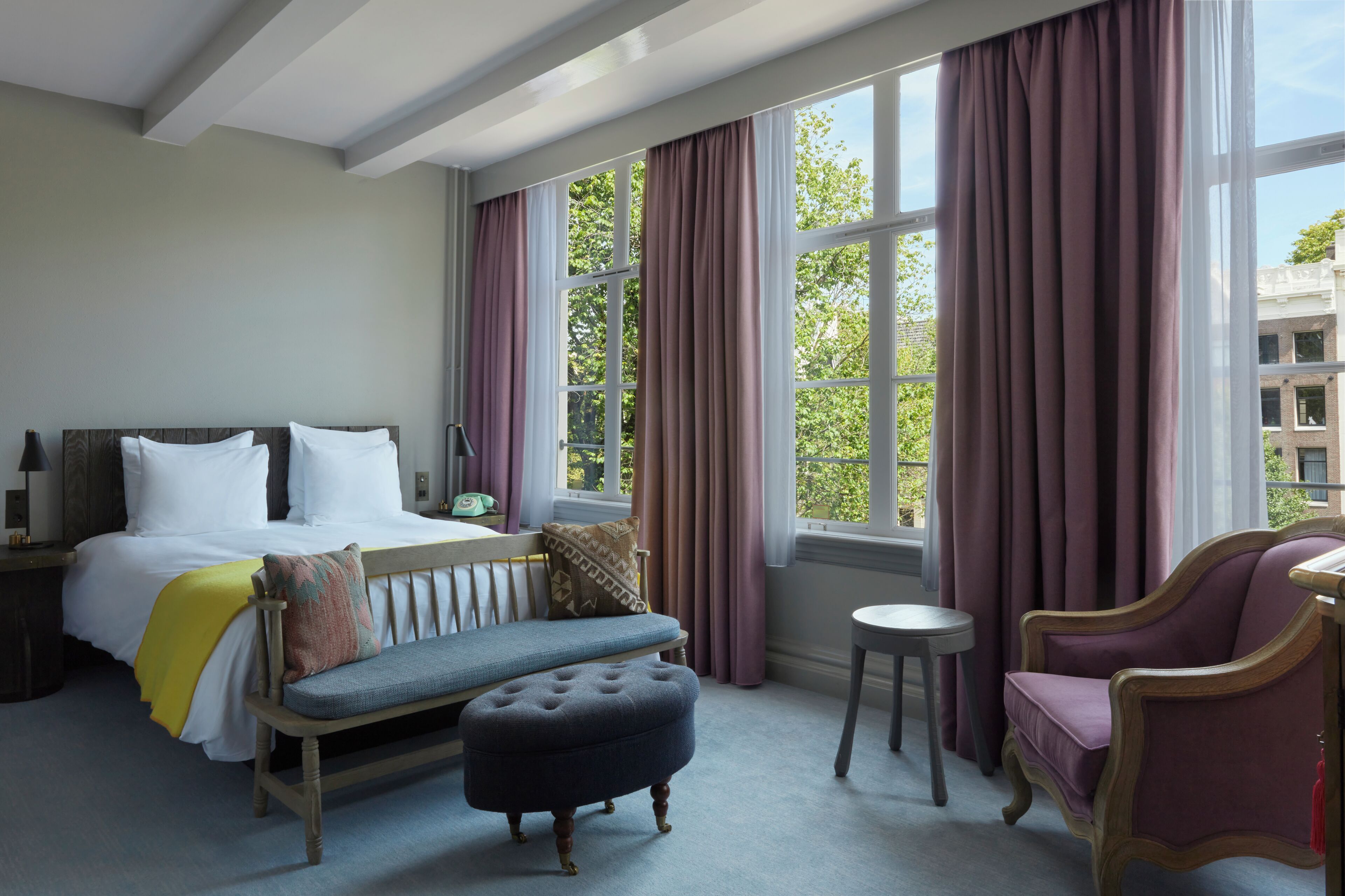 Une chambre d’hôtel avec un lit, un banc, un pouf, une chaise et une vue sur les arbres, visible à travers de grandes fenêtres.