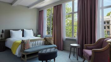 Une chambre d’hôtel avec un lit, un banc, un pouf, une chaise et une vue sur les arbres, visible à travers de grandes fenêtres.