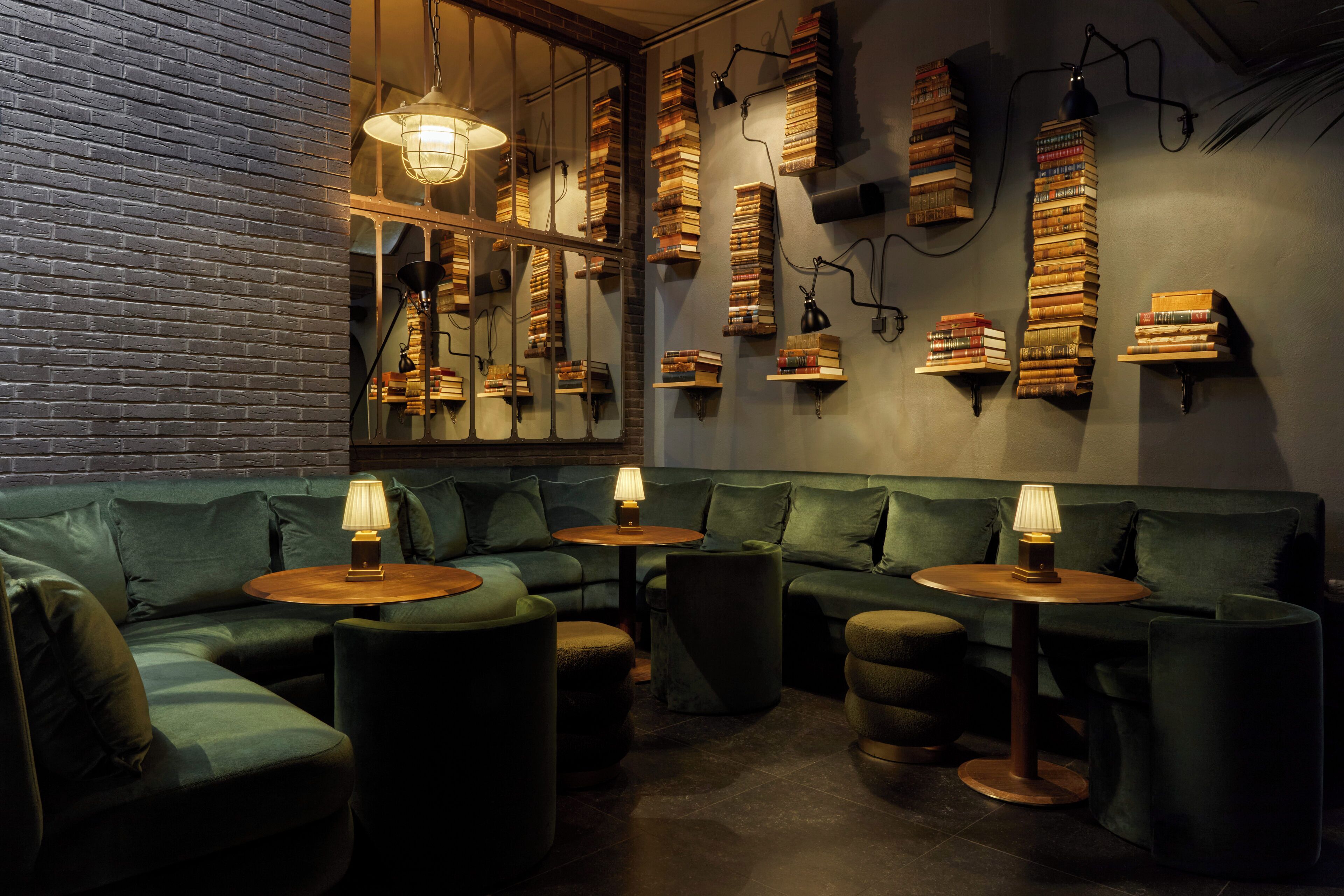 A cozy lounge area with green upholstered seating, small tables with lamps, and bookshelves filled with books.