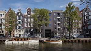 A canal with two boats, a row of historic buildings with uniform windows and facades, and a clear blue sky. - Pulitzer Amsterdam (Amsterdam)