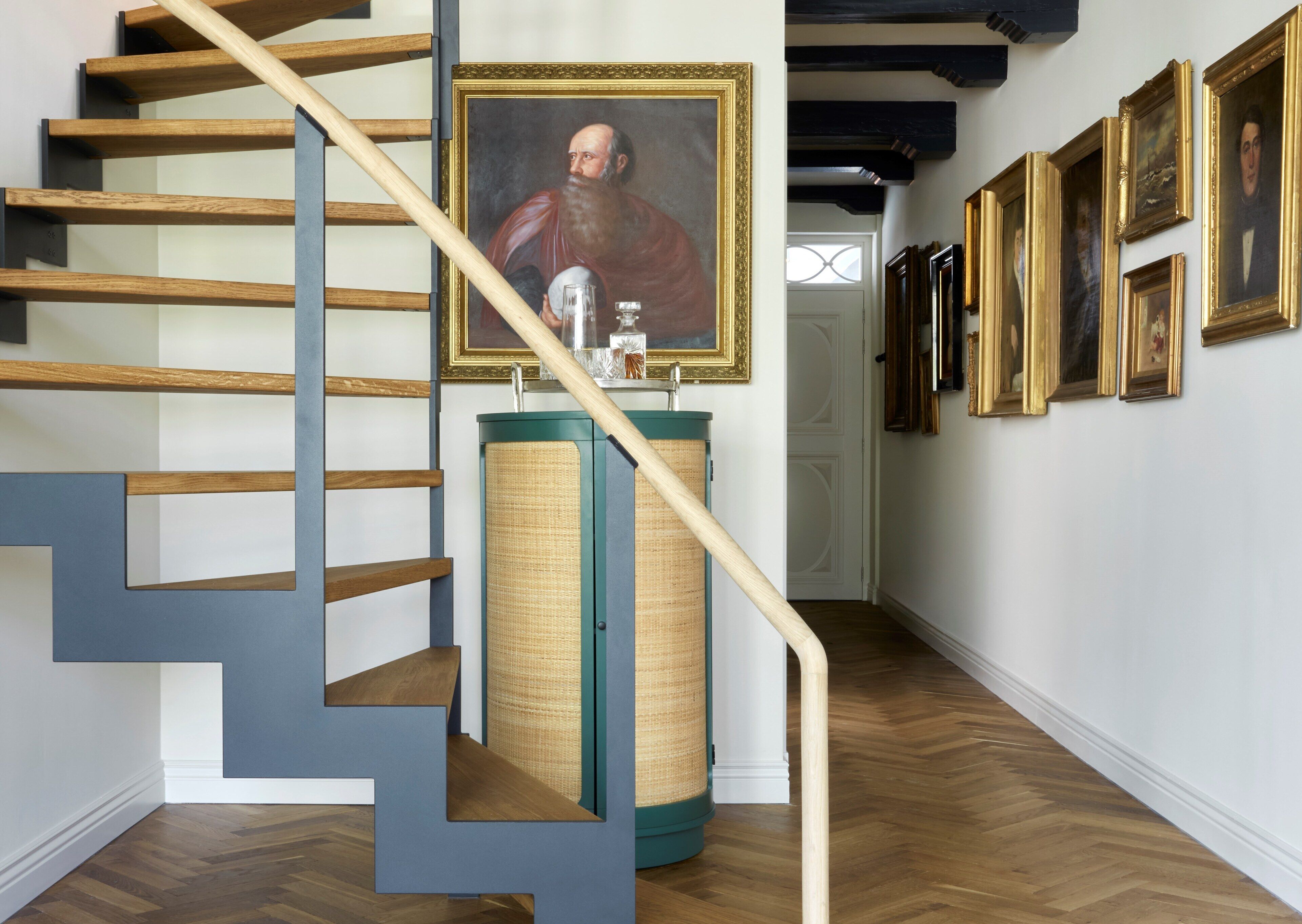 Una escalera con escalones de madera y barandilla azul, que conduce a una habitación con un cuadro grande y un armario verde.