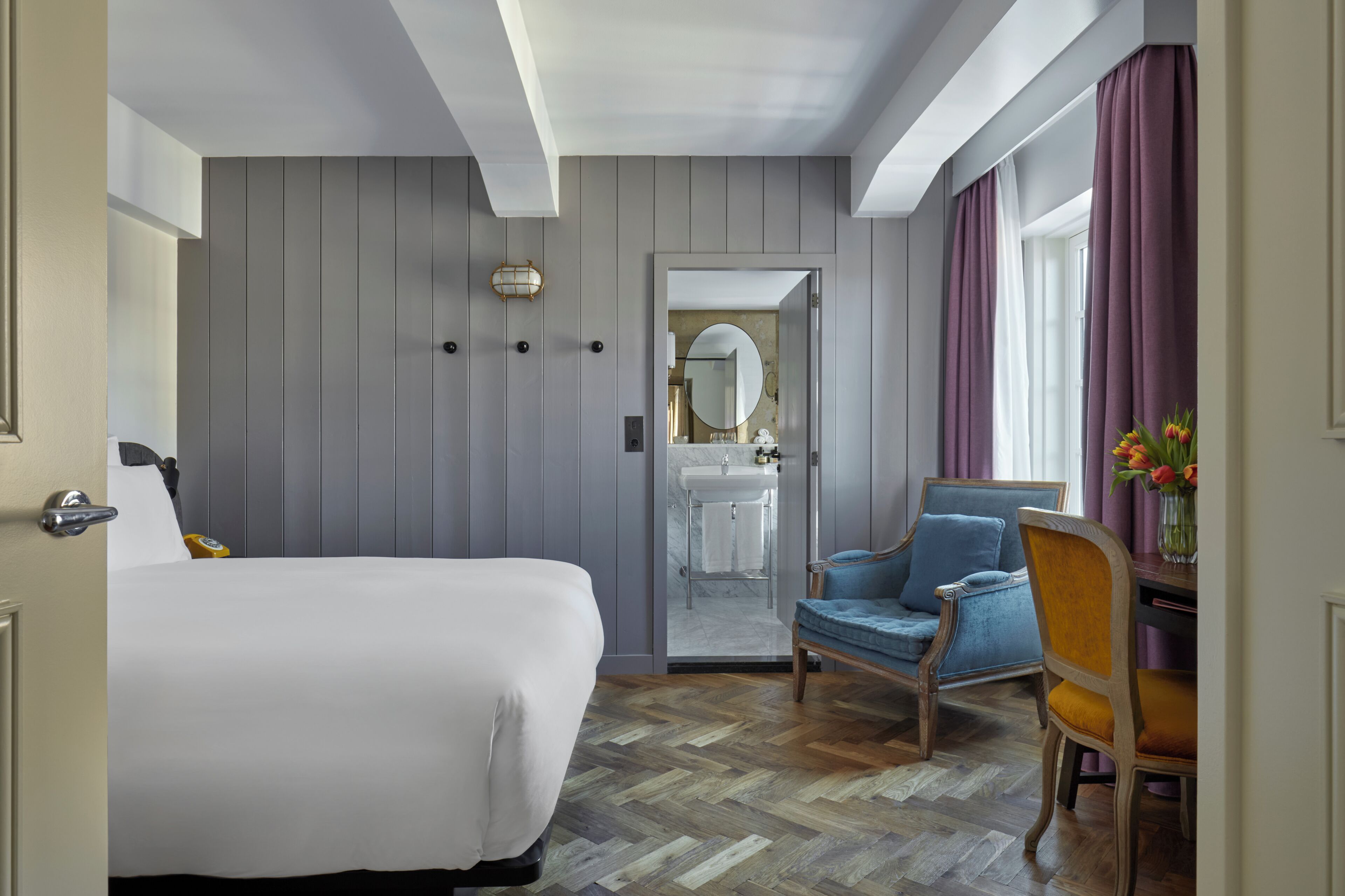 A hotel room with a bed, a chair, a desk, a mirror, and a bathroom with a sink and shower.