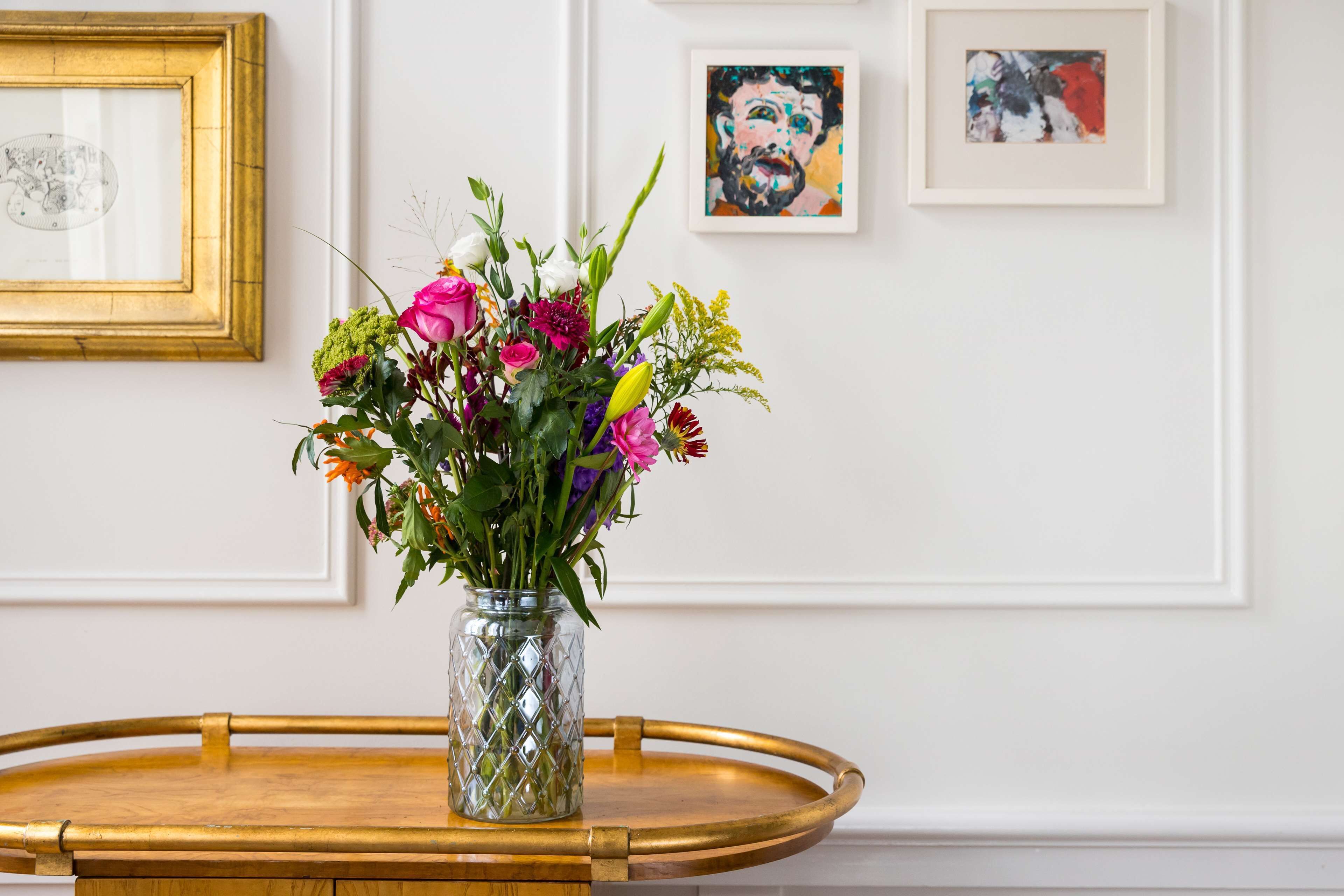 Une pièce avec une petite table en bois, un vase en verre rempli de fleurs et des œuvres d’art encadrées accrochées au mur.